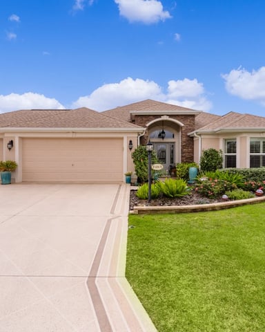 Single-story beige stucco home with brick accents, three-car garage, manicured lawn, and landscaped front garden under blue sky.