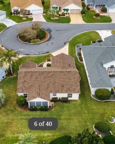 Aerial view of single-story home with brown roof, white trim, and screened porch surrounded by manicured lawn and palm trees in residential community.