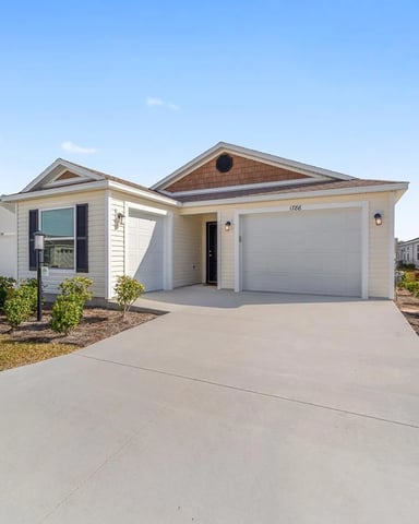 Single-story beige house with white garage door, pitched roof, and landscaped front yard in residential community