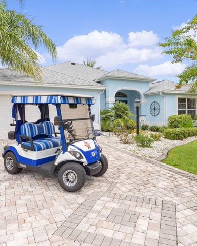 Blue and white golf cart parked on driveway in front of light blue single-story home with palm trees and manicured landscaping