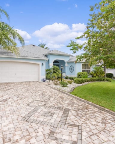 Light blue single-story home with white garage, paver driveway, palm trees, and manicured lawn