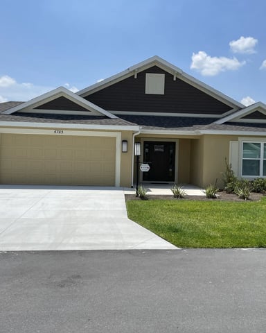 Single-story tan house with dark roof, two-car garage, black front door, and manicured lawn under blue sky