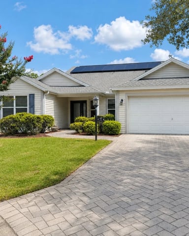 Single-story white house with solar panels, two-car garage, brick driveway, and manicured landscaping with flowering shrub.
